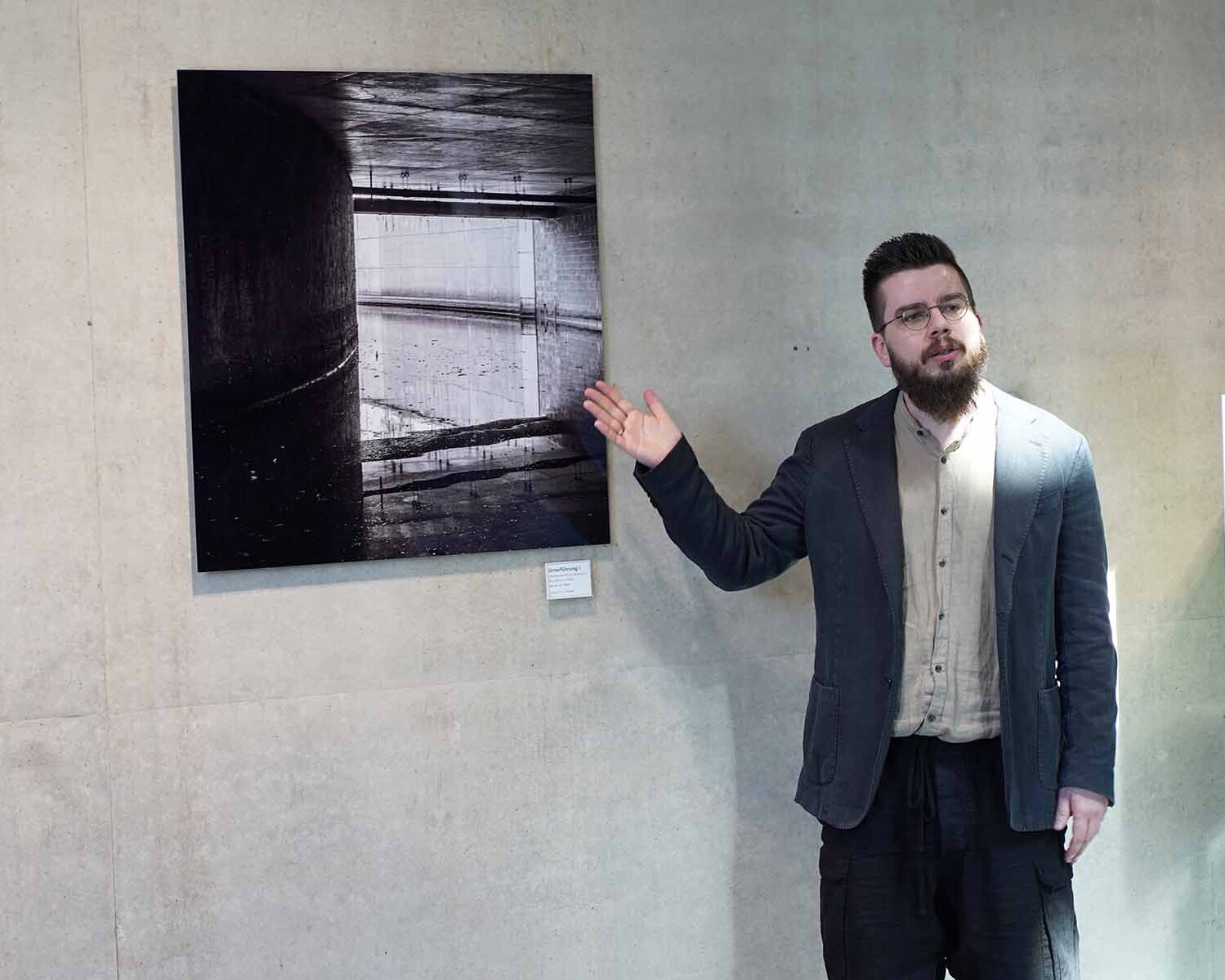 Der Künstler steht vor einer Fotografie an der Wand und zeigt mit der Hand darauf.