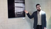 Der Künstler steht vor einer Fotografie an der Wand und zeigt mit der Hand darauf.