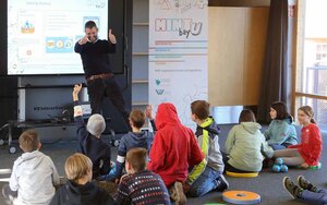 Ein Mann hebt beide Daumen und lächelt die vor ihm auf dem Boden sitzenden Kinder an.