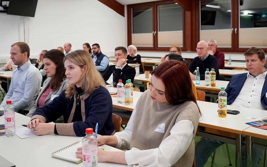 Teilnehmende sitzen an Tischen im Hörsaal, eine Frau schreibt mit