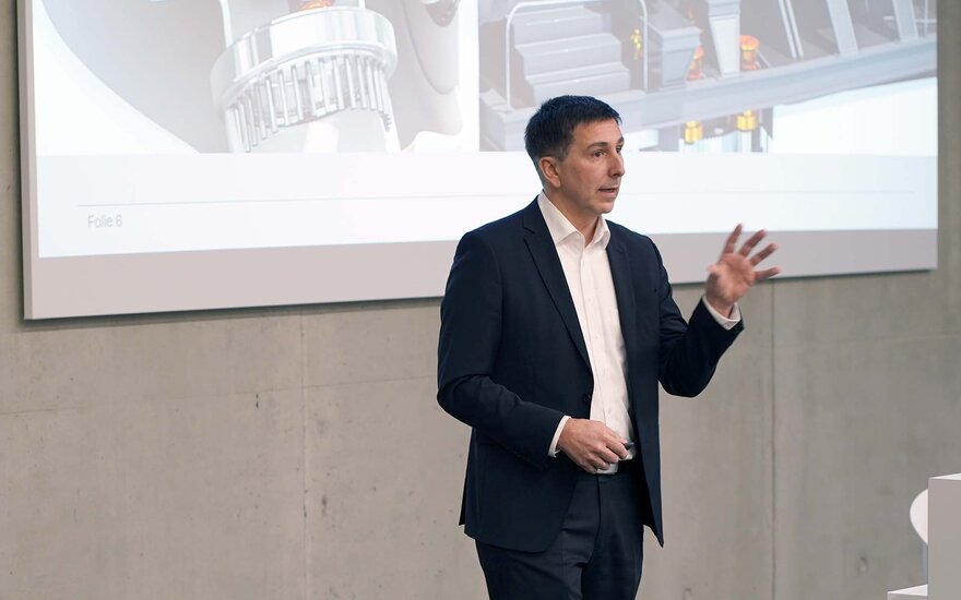 Mann mit kurzen dunklen Haaren im schwarzen Anzug spricht gestikulierend vor Leinwand