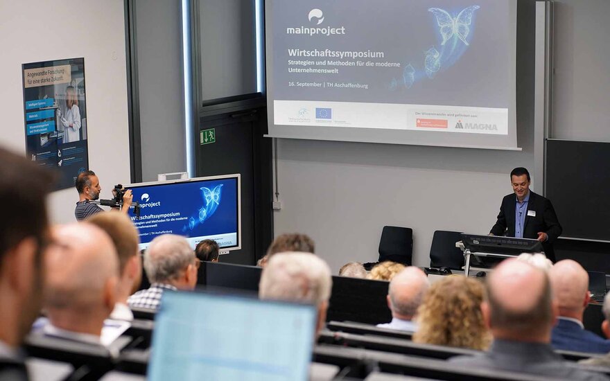 Vortragender vor Screen mit Beschriftung zum Symposium mit Teilnehmenden im Hörsaal 