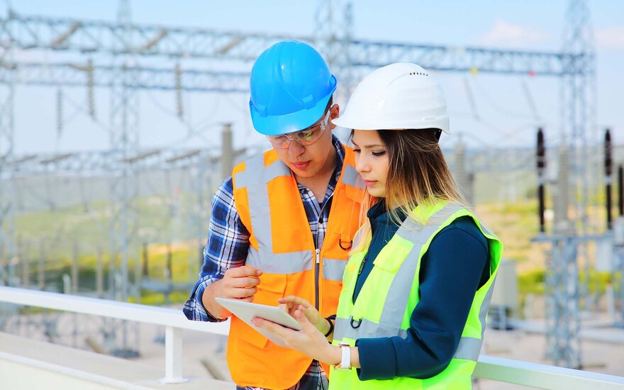 Eine Frau und ein Mann mit Bauhelmen und Warnwesten an einem Tablet in einem Umspannwerk