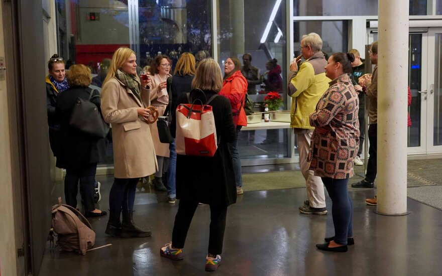 Eine Gruppe von Personen steht im Foyer zusammen. Die Personen unterhalten sich bei einem kleinen Imbiss.