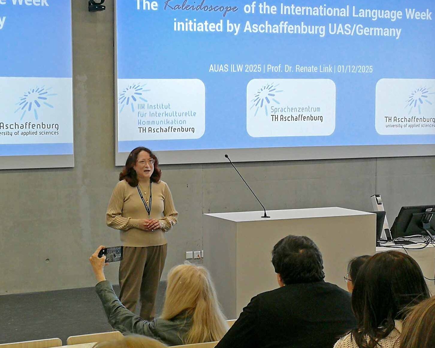 Frau mit roten schulterlangen Haaren und Brille spricht vor cyanblauer Präsentation zum Publikum