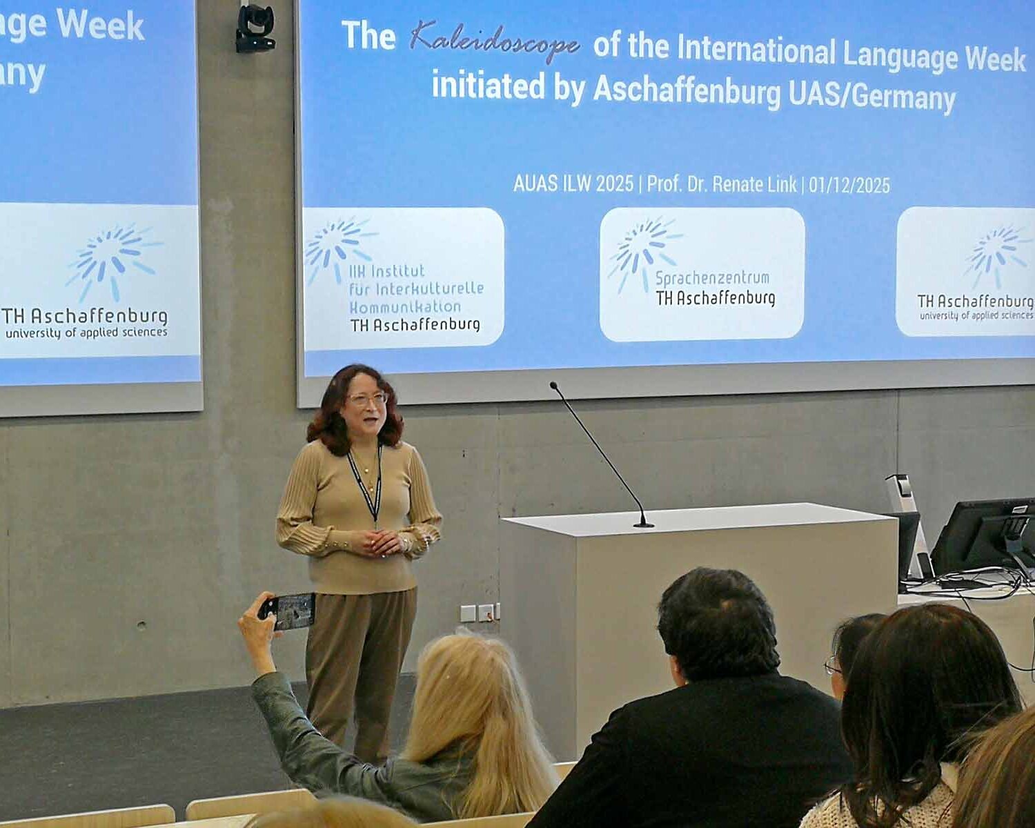 Frau mit roten schulterlangen Haaren und Brille spricht vor cyanblauer Präsentation zum Publikum