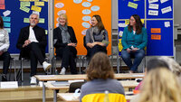 Eine Gruppe von 5 Personen sitzt vor sitzendem Publikum