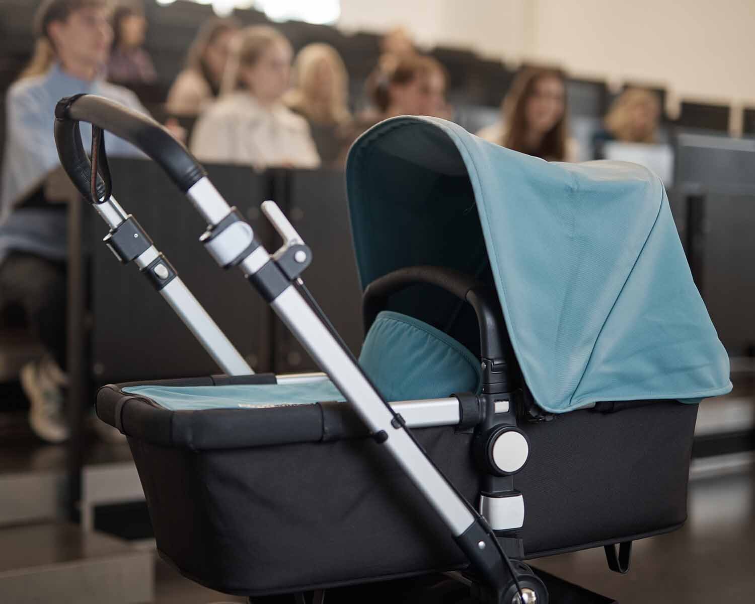 Kinderwagen steht im Hörsaal vor ansteigenden Reihen mit Studierenden