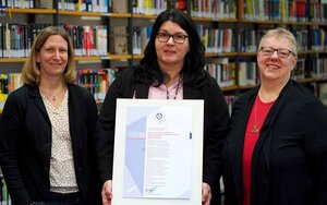 Drei Frauen stehen nebeneinander vor einem Bücherregal, die Frau in der Mitte hält den Ausdruck eines Zertifikats in der Hand
