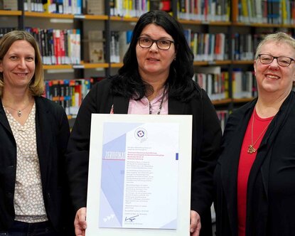 Drei Frauen stehen nebeneinander vor einem Bücherregal, die Frau in der Mitte hält den Ausdruck eines Zertifikats in der Hand