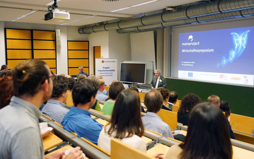 Personen sitzen im ansteigenden Hörsaal und schauen auf blaue Leinwand mit der Aufschrift mainproject Wirtschaftssymposium und den vortragenden Professor