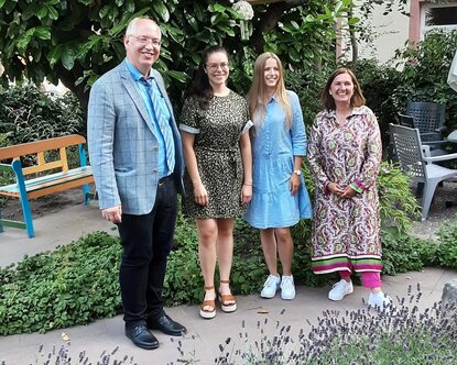 Zwei junge Frauen in Sommerkleidern stehen in einem Garten mit einem Professor und einer Frau mittleren Alters