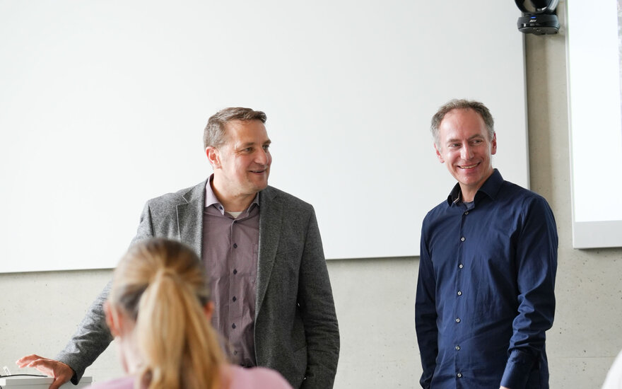 Zwei Professoren stehen im Innenraum vor einer Projektionsfläche und sind in ein Gespräch eingebunden; im Vordergrund ist am unteren Bildrand eine weitere Person teilweise sichtbar.