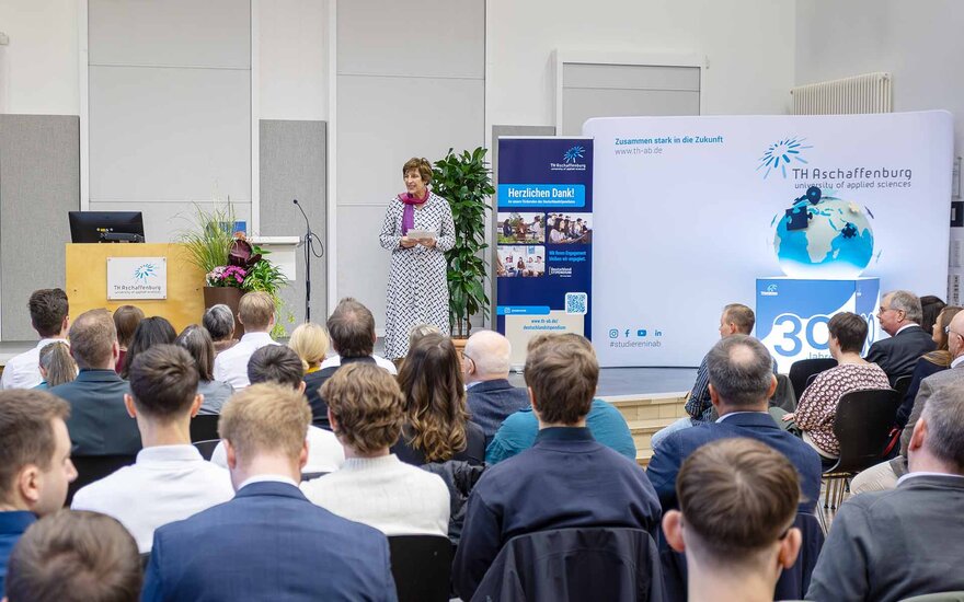 Frau mit kurzen dunklen Haaren mit Mikrophon steht auf der Bühne der Aula vor sitzendem Publikum