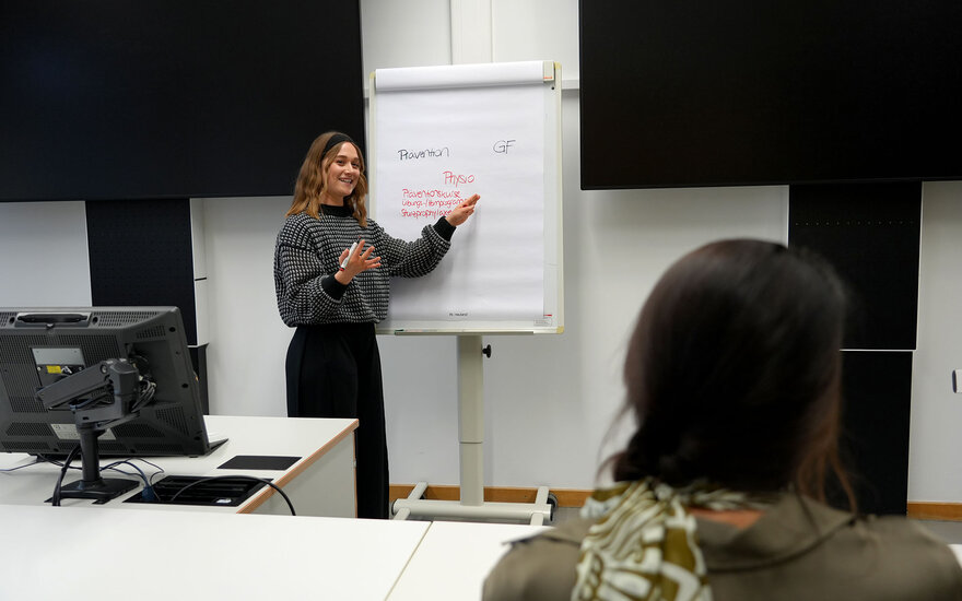 Eine Studentin skizziert Themen aus ihrem Berufsalltag an einem Whiteboard, während eine andere Studentin ihr zuhört
