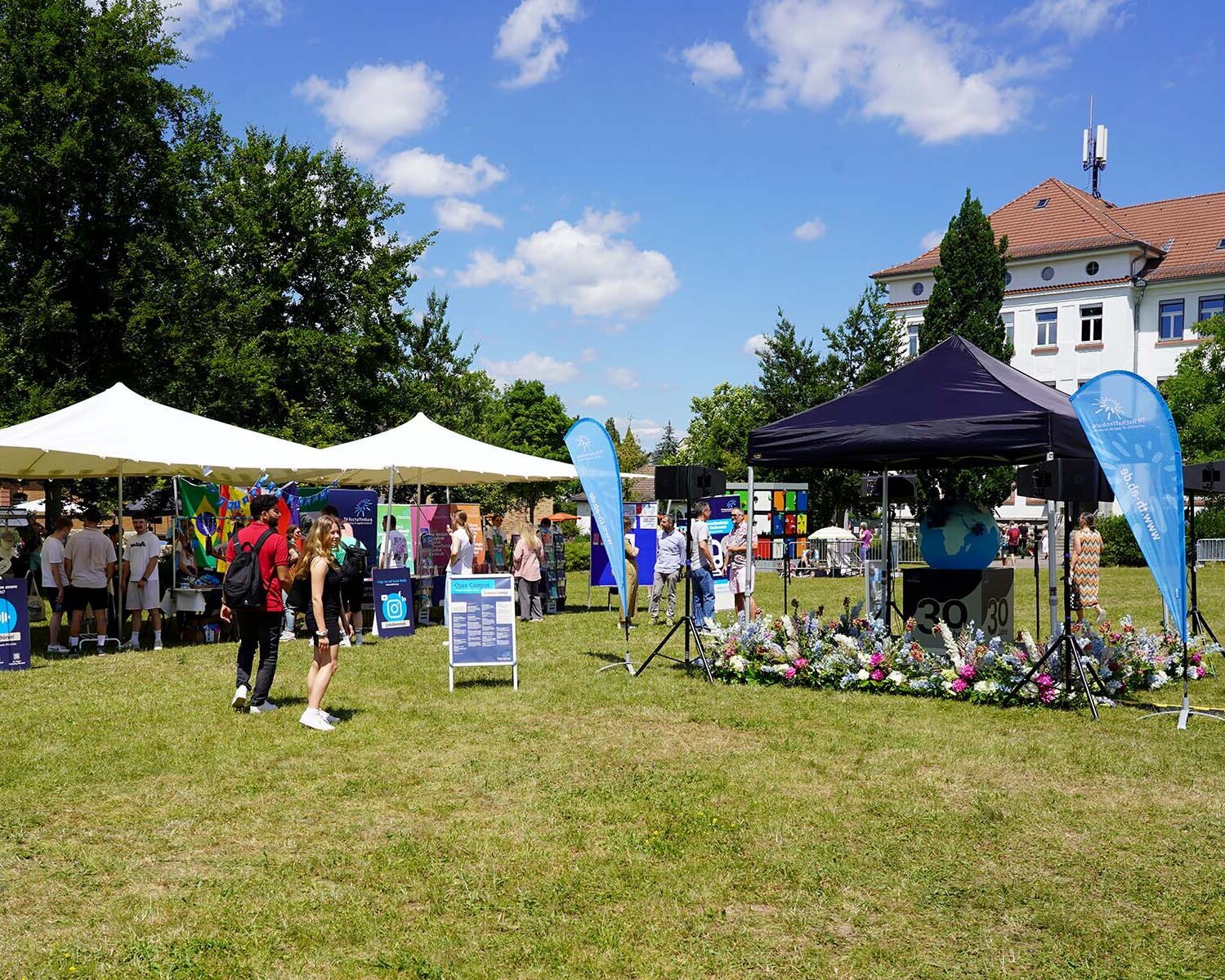 Campuswiese mit Menschen an Infoständen und weißen Zelten im Hintergrund sowie 30-Jahre-Keyvisual auf blau überdachter Bühne