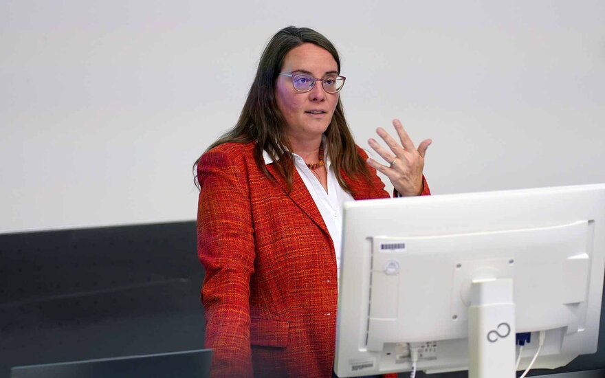 Eine Frau mit langen braunen Haaren, einer Brille und einem roten Blazer steht hinter einen Computerbildschirm und spricht zum Publikum