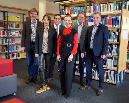 Zwei Professorinnen und vier Professoren stehen nebeneinander vor Bücherregalen in der Bibliothek und lächeln in die Kamera