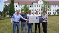 Zwei Professoren, zwei Mitarbeiter von Buderus und die Präsidentin halten auf der Campuswiese vor dem weißen Gebäude eine graue Tafel mit Firmenlogo und Bezeichnung der Wärmepumpe hoch