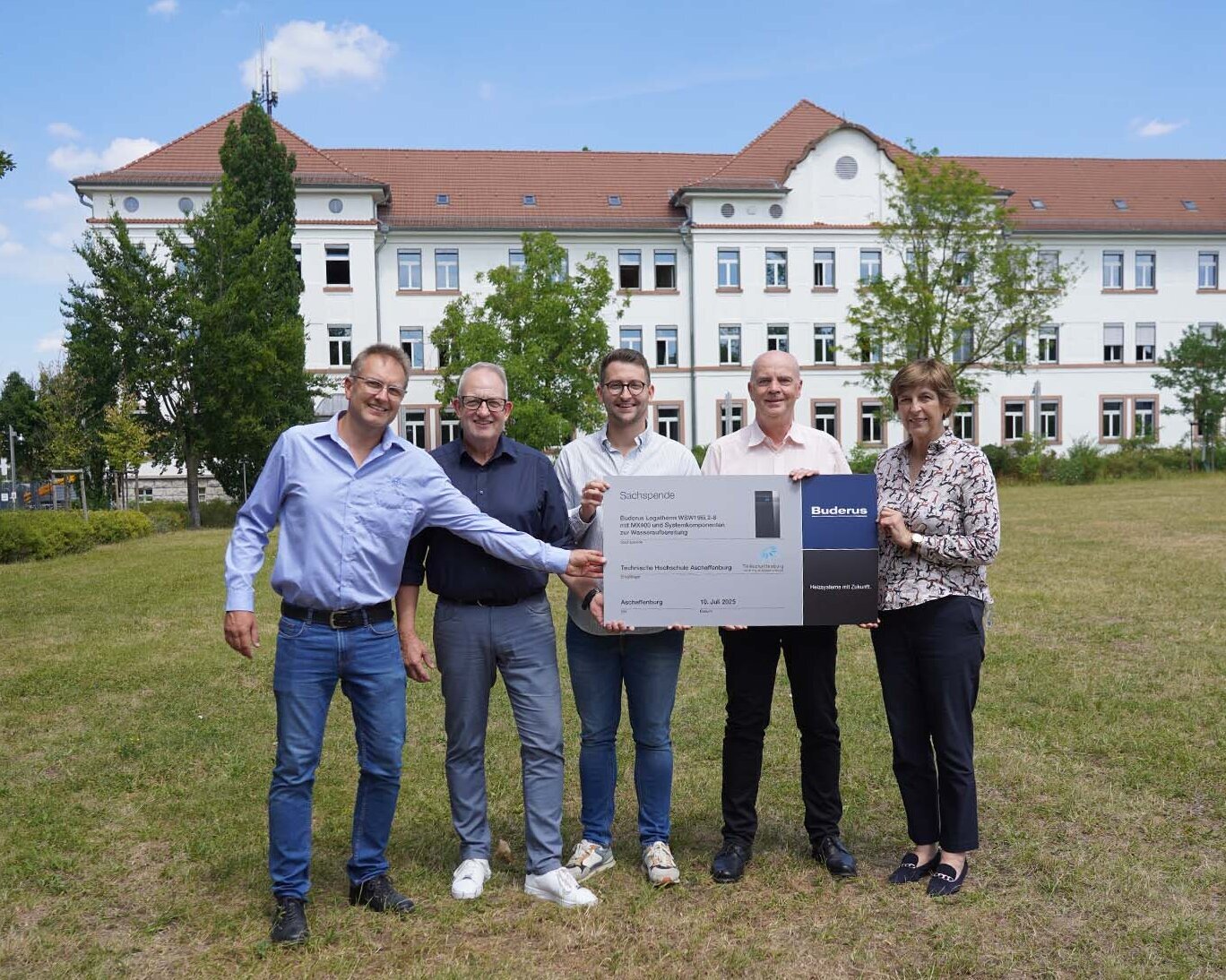 Zwei Professoren, zwei Mitarbeiter von Buderus und die Präsidentin halten auf der Campuswiese vor dem weißen Gebäude eine graue Tafel mit Firmenlogo und Bezeichnung der Wärmepumpe hoch