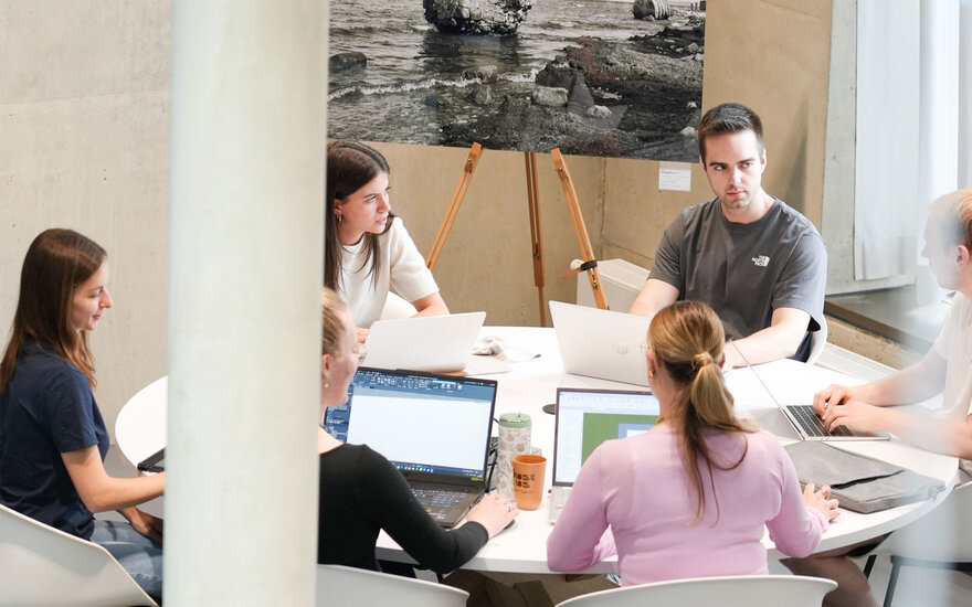 Mehrere Studierende sitzen gemeinsam an einem runden Tisch und arbeiten an offenen Laptops; auf dem Tisch stehen ein Getränkebecher und Unterlagen, während im Hintergrund ein auf einer Staffelei platziertes Bild sowie helle Wände und Fenster den Raum ergänzen.