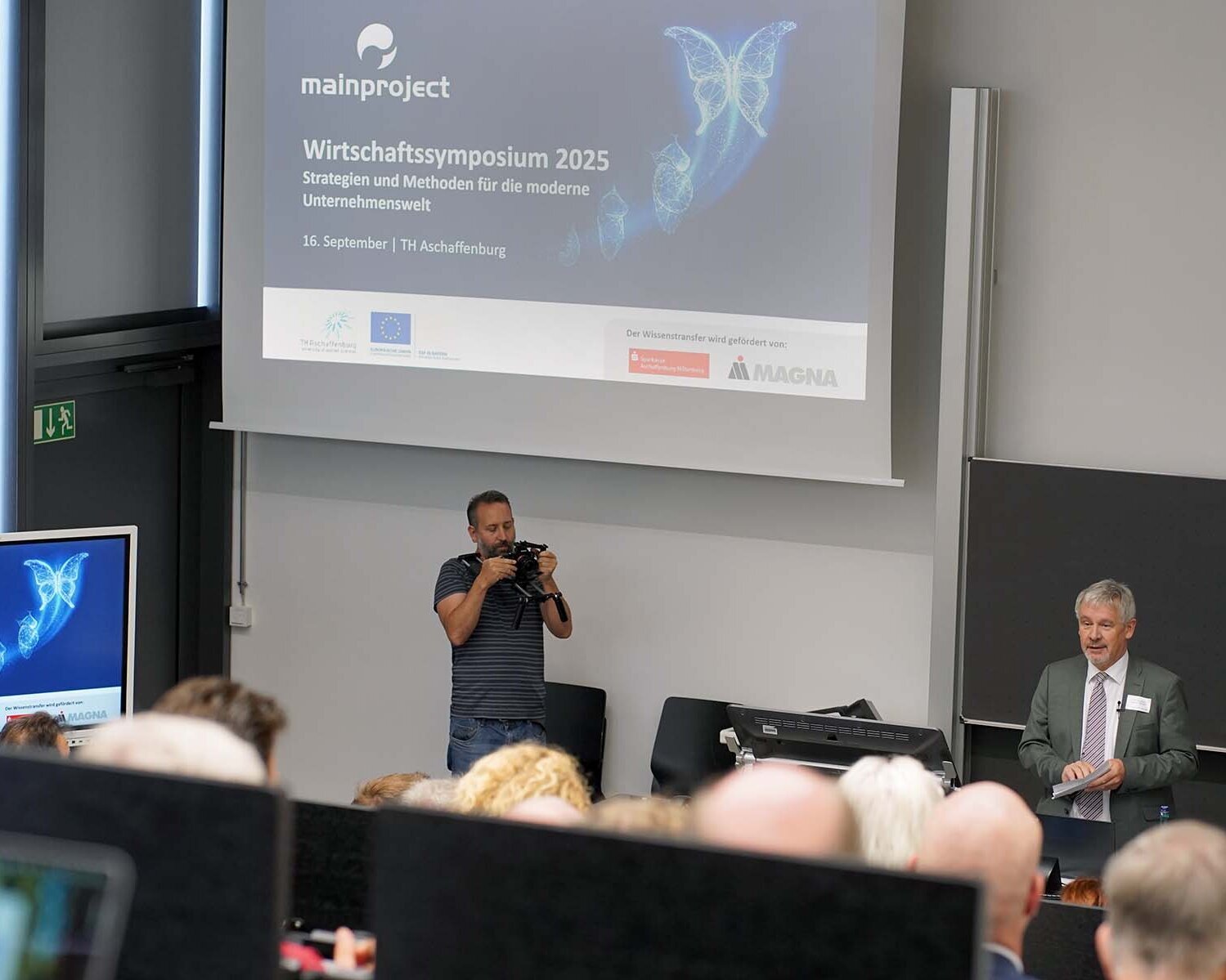Professor Hofmann spricht vor Teilnehmenden im Hörsaal mit Beamerbild zum Wirtschaftssymposium im Hintergrund.
