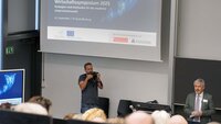 Professor Hofmann spricht vor Teilnehmenden im Hörsaal mit Beamerbild zum Wirtschaftssymposium im Hintergrund.