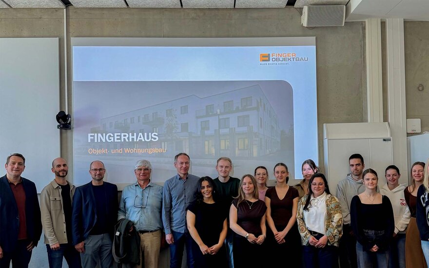 Gruppe von etwa 15 Personen steht in einem Vorlesungsraum vor einer projizierten Präsentationsfolie mit der Überschrift „FINGERHAUS Objekt- und Wohnungsbau“; im Hintergrund ist eine Visualisierung eines Gebäudekomplexes zu sehen.