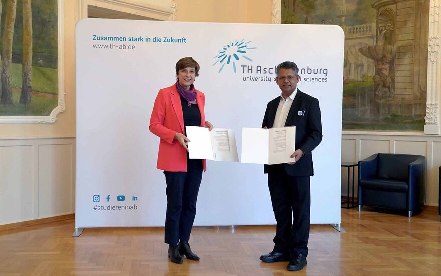 Frau in rotem Blazer und Mann im schwarzen Anzug halten unterschriebene Dokumente in der Hand