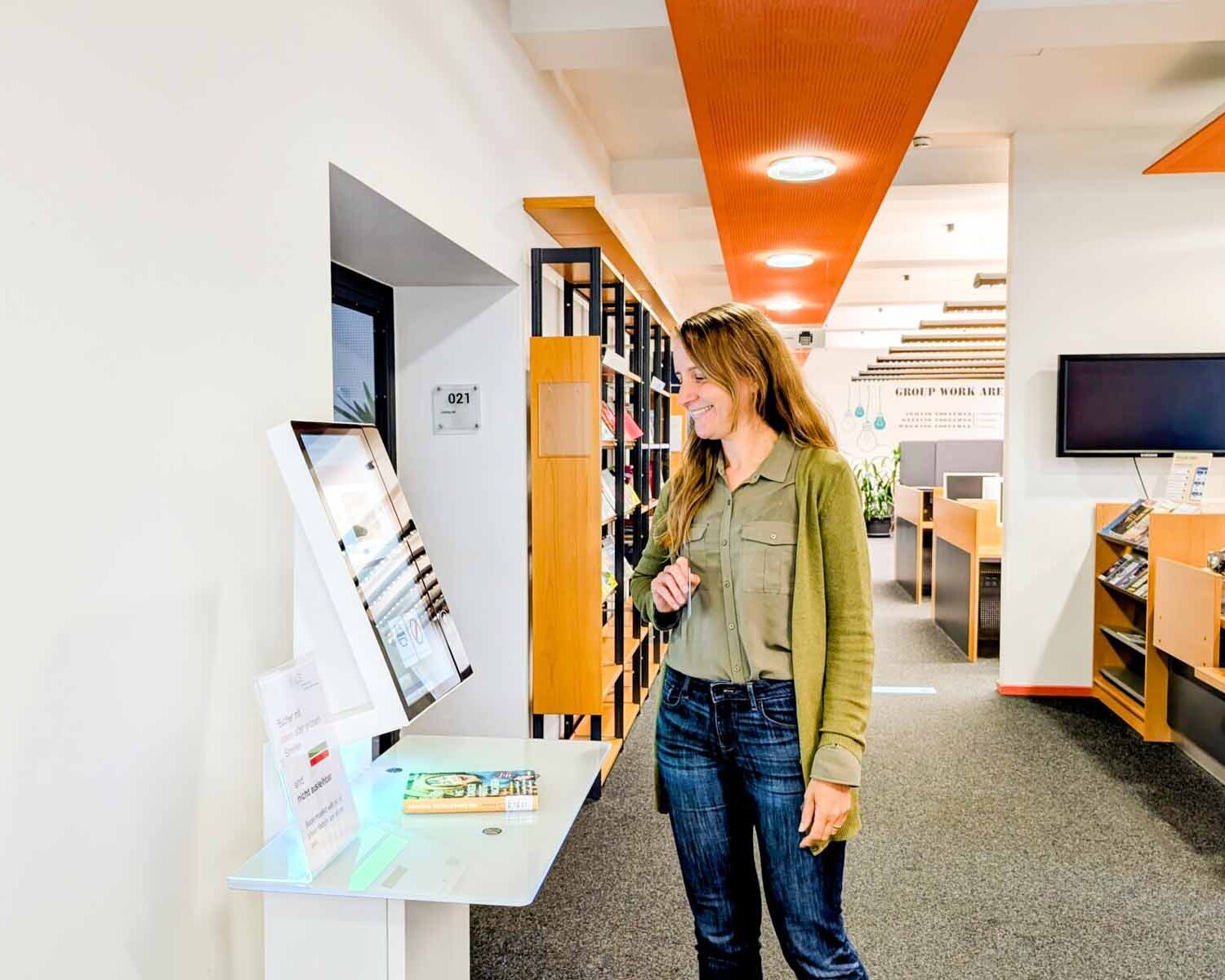 Frau mit langen blonden Haaren blickt auf großen Bildschirm vor Bücherregalen