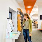 Frau mit langen blonden Haaren blickt auf großen Bildschirm vor Bücherregalen