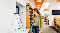 Frau mit langen blonden Haaren blickt auf großen Bildschirm vor Bücherregalen