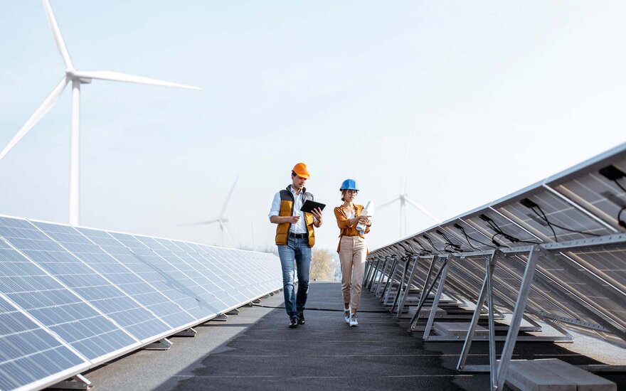 Ein Mann und eine Frau mit Helmen laufen über einen Solarpark. Im Hintegrund sind Windräder