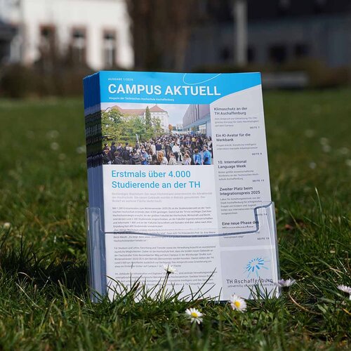 Hochschulmagazine im Prospektständer auf Campuswiese vor weißem und grauem Gebäude