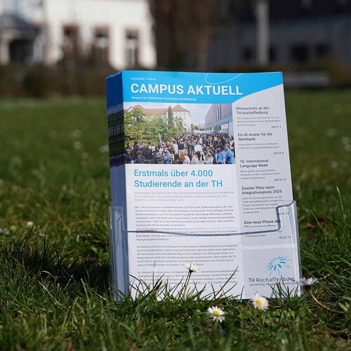 Hochschulmagazine im Prospektständer auf Campuswiese vor weißem und grauem Gebäude