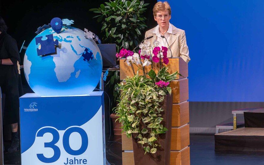 Frau mit kurzen blonden Haaren und Brille im rosa Blazer am Rednerpult