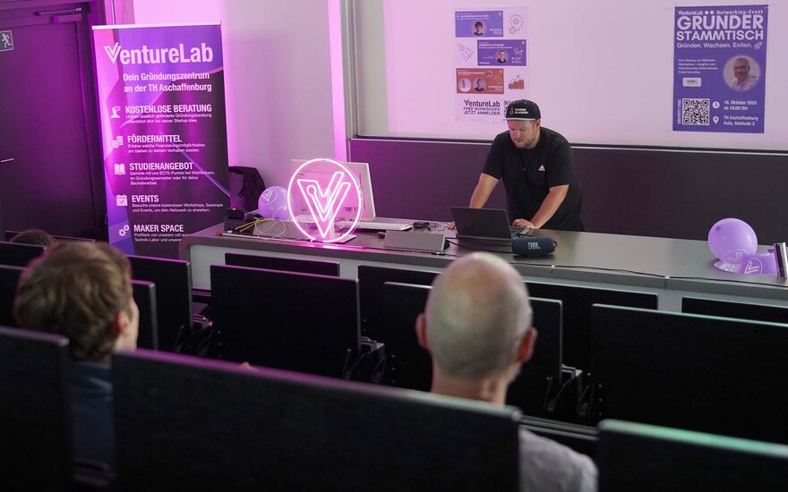 Ein junger Mann im schwarzen T-Shirt hält einen Vortrag im Hörsaal mit verschiedenen Personen von hinten im Vordergrund, mehrere Poster in Pink und Lila sowie ein pink beleuchtetes V als Logo des VentureLabs sind auf und um den Tisch dekoriert