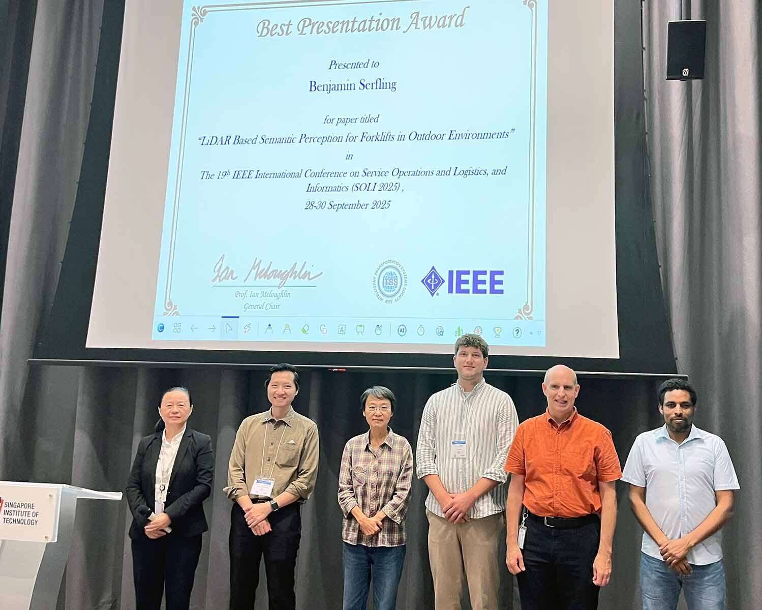 Gruppenbild mit sechs Männern und Frauen. Sie stehen vor einer Leinwand, auf der ein Bild der Urkunde vom Best Presentation Award von Benjamin Serfling abgebildet ist. 