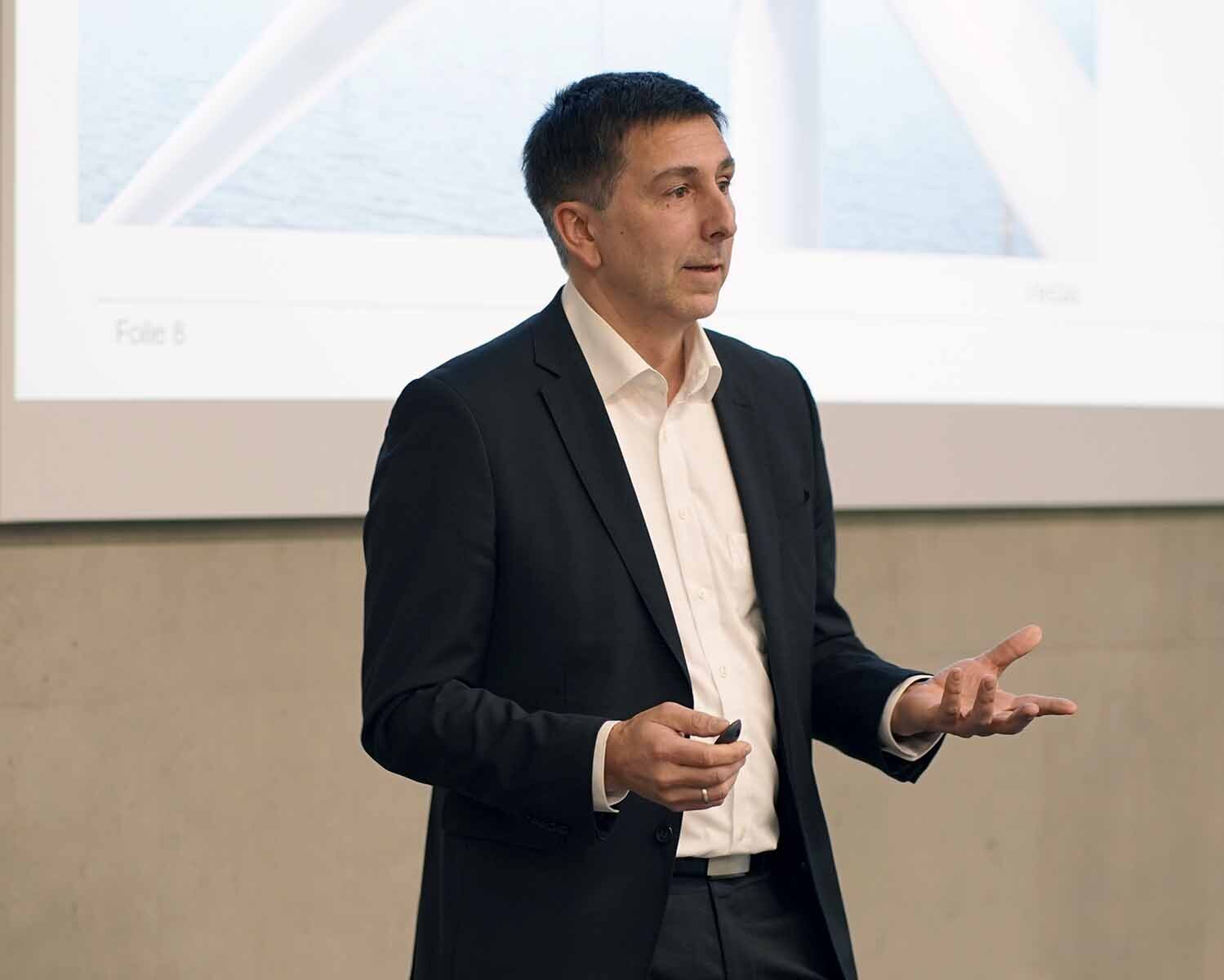 Mann mit kurzen dunklen Haaren im schwarzen Anzug spricht gestikulierend vor Leinwand