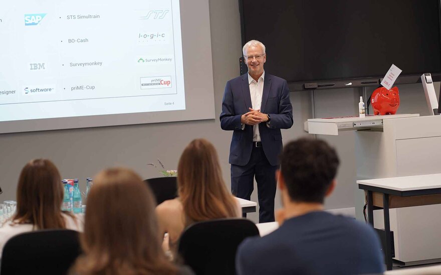 Professor spricht vor Screen lächelnd zu den Studierenden