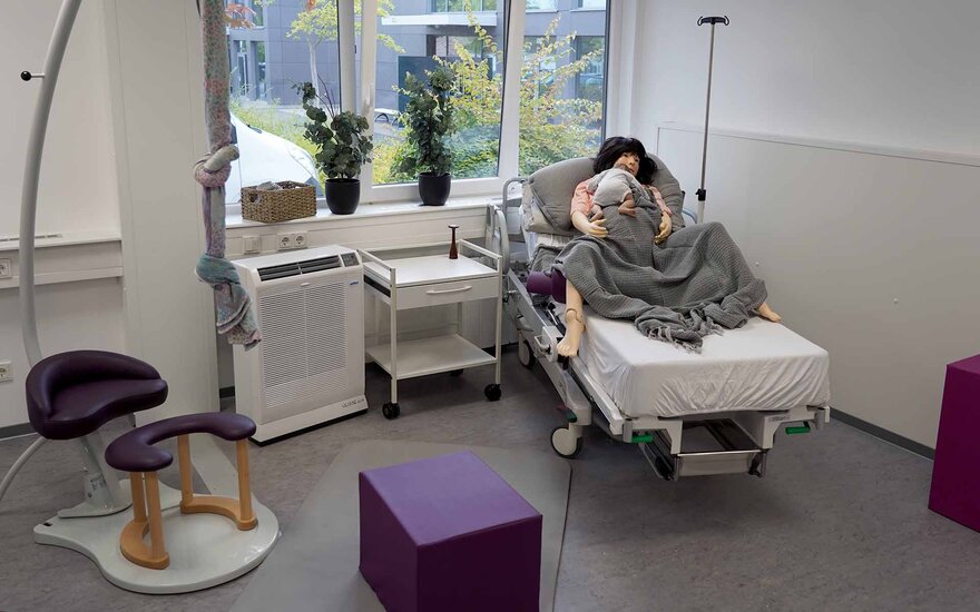 Ein Raum mit einem Bett, in dem eine Simulationspuppe in Größe einer erwachsenen Frau mit einem Baby im Arm liegt