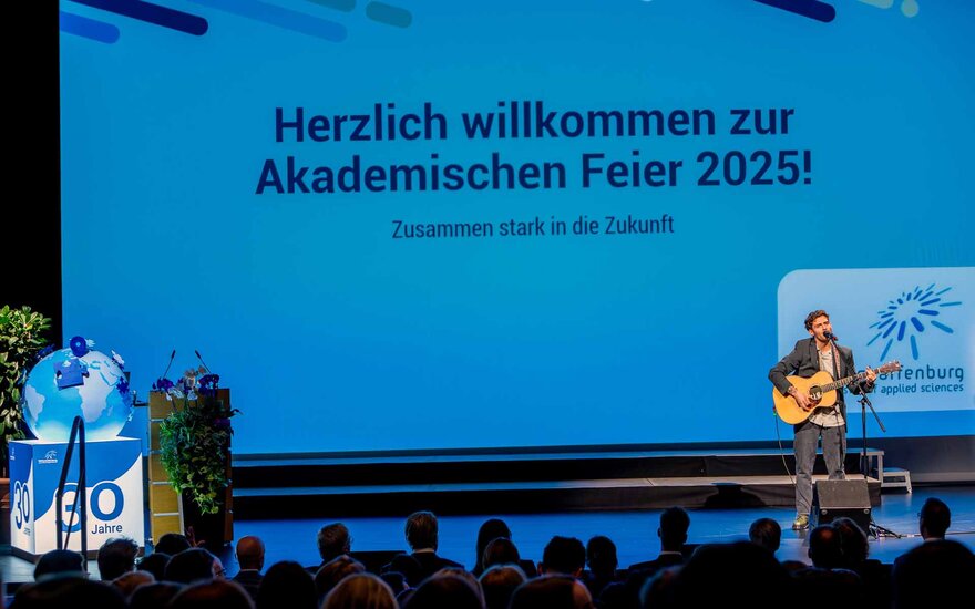 Musiker mit Gitarre vor blauem Beamerbild mit Herzlich willkommen und TH-Logo