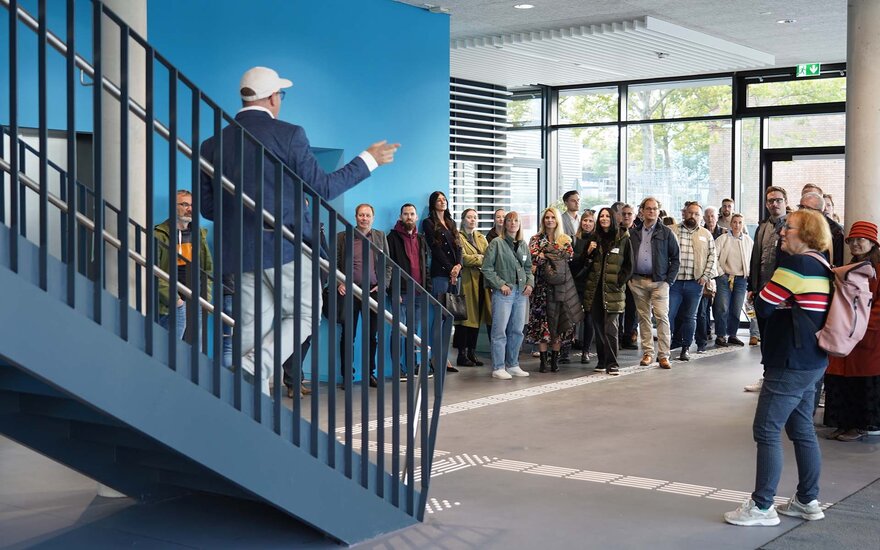 Mann mit Kappe und blauem Jackett von hinten, der auf einer Treppe steht und einer großen Gruppe von Personen etwas erklärt