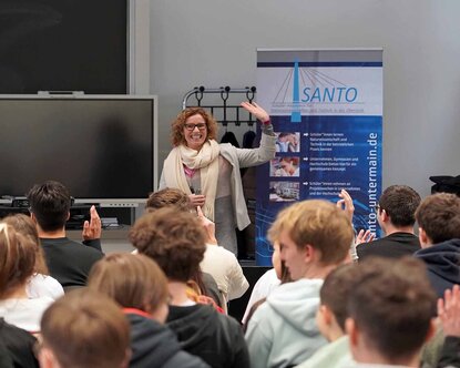 Professorin McNamara steht lächelnd vor den sitzenden Schülerinnen und Schülern und hebt die Hand. Einige Schüler melden sich. Im Hintergrund ist ein blau-weißes SANTO-Roll-up zu sehen.