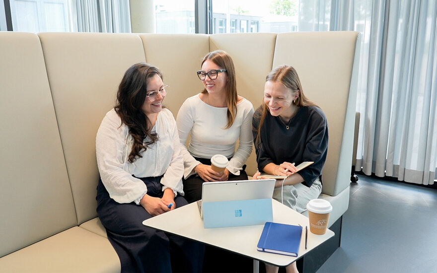 Drei Studentinnen sitzen an einem Tisch in einem modernen Aufenthaltsraum und arbeiten am Laptop.
