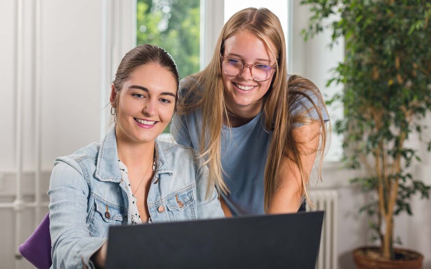 Zwei lachende WR-Studentinnen vor einem Laptop. Im Hintergrund eine Pflanze.