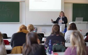 Eine Frau mit kurzen lockigen blonden Haaren spricht vor einer Gruppe von sitzenden Frauen in einem Hörsaal