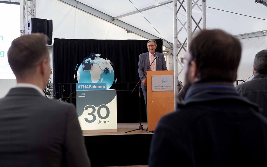 Älterer Mann in hellgrauem Anzug, mit grauen Haaren und Brille steht am Rednerpult aus Holz mit TH-Logo darauf, links daneben steht ein beleuchteter großer Würfel mit einem blauen 30-Jahre-Schriftzug und einem blau-grauen Globus obendrauf, zwei Männer mit Blick zur Bühne sind von hinten zu sehen
