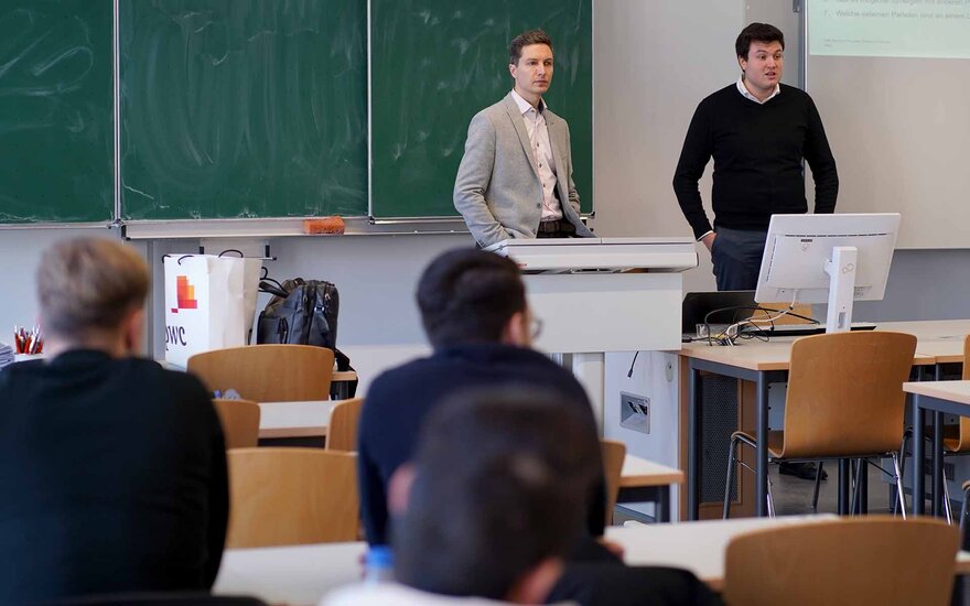 Zwei Männer stehen nebeneinander vor einer grünen Tafel und sprechen zu den Studierenden