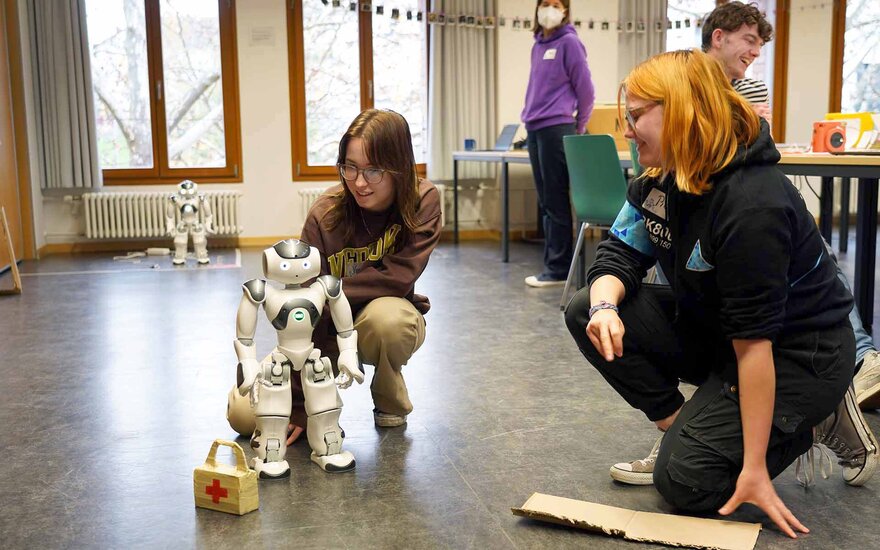 Zwei Schülerinnen hocken auf dem Boden neben dem NAO-Roboter und zeigen, wie er sich bewegt.